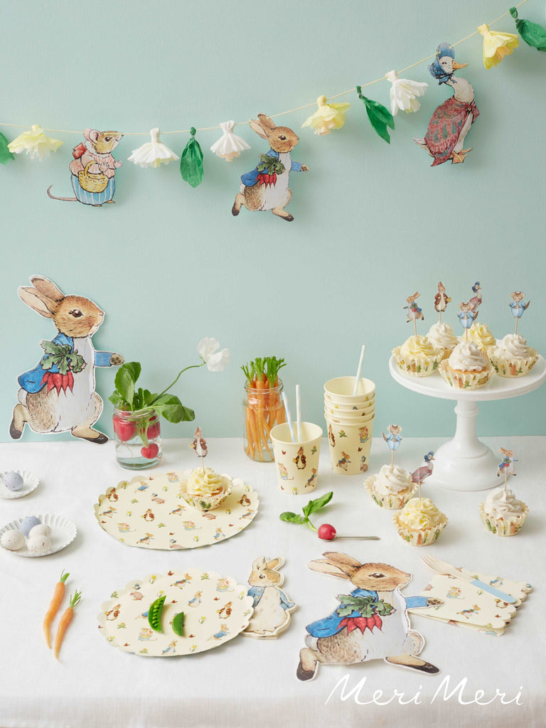 Meri Meri Peter Rabbit party supplies set a beautiful table. The table is set with cups, napkins and plates that feature all the Beatrix Potter animals. There are cupcakes on a whtie cake stand with carrots and radishes in jars on the table.