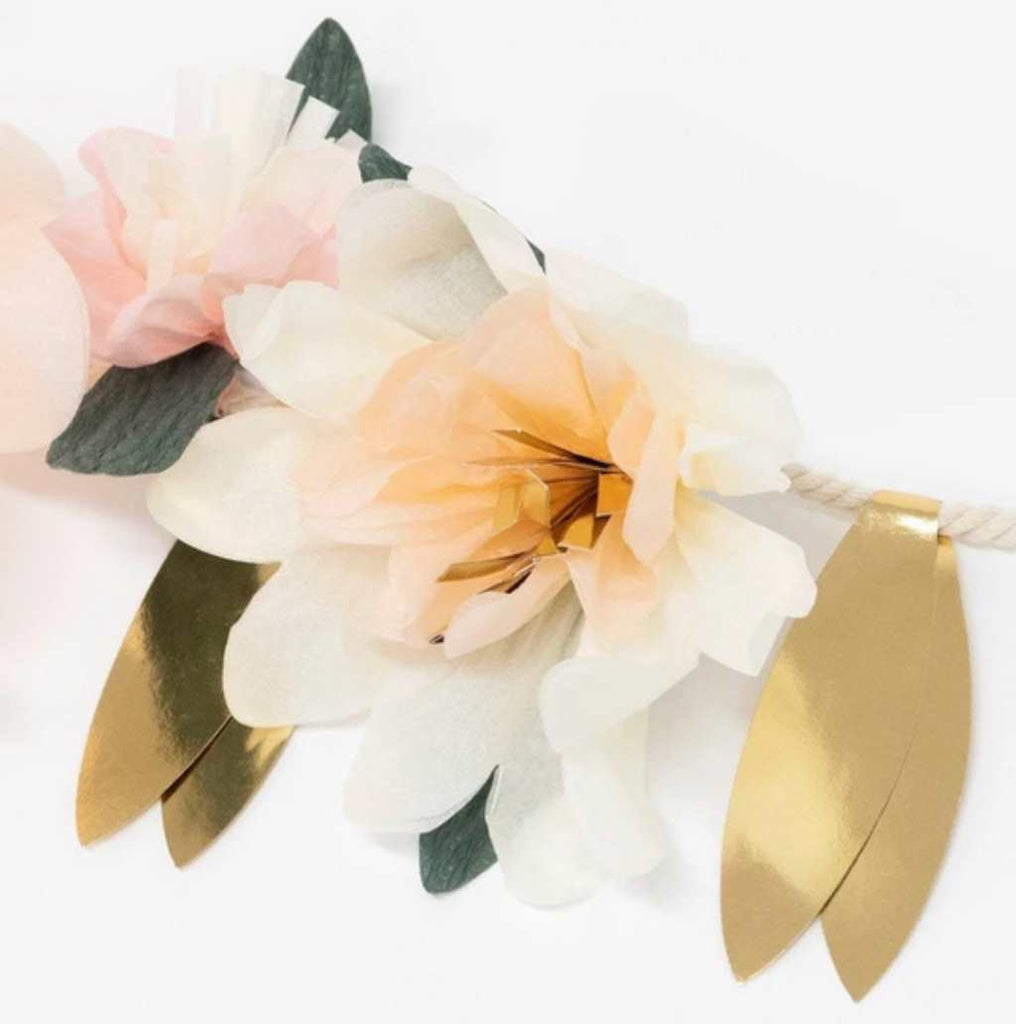 cream tissue paper flower hanging on a rope with gold paper leaves on either side. There is an out of focus pink tissue flower to the left of the cream flower.