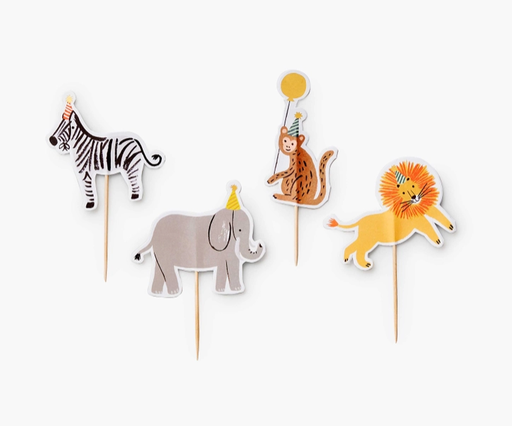 cupcake toppers on toothpicks laying in a row a zebra with a party hat, an elephant with a party hat, a monkey wearing a party hat holding a balloon and a lion with a party hat.